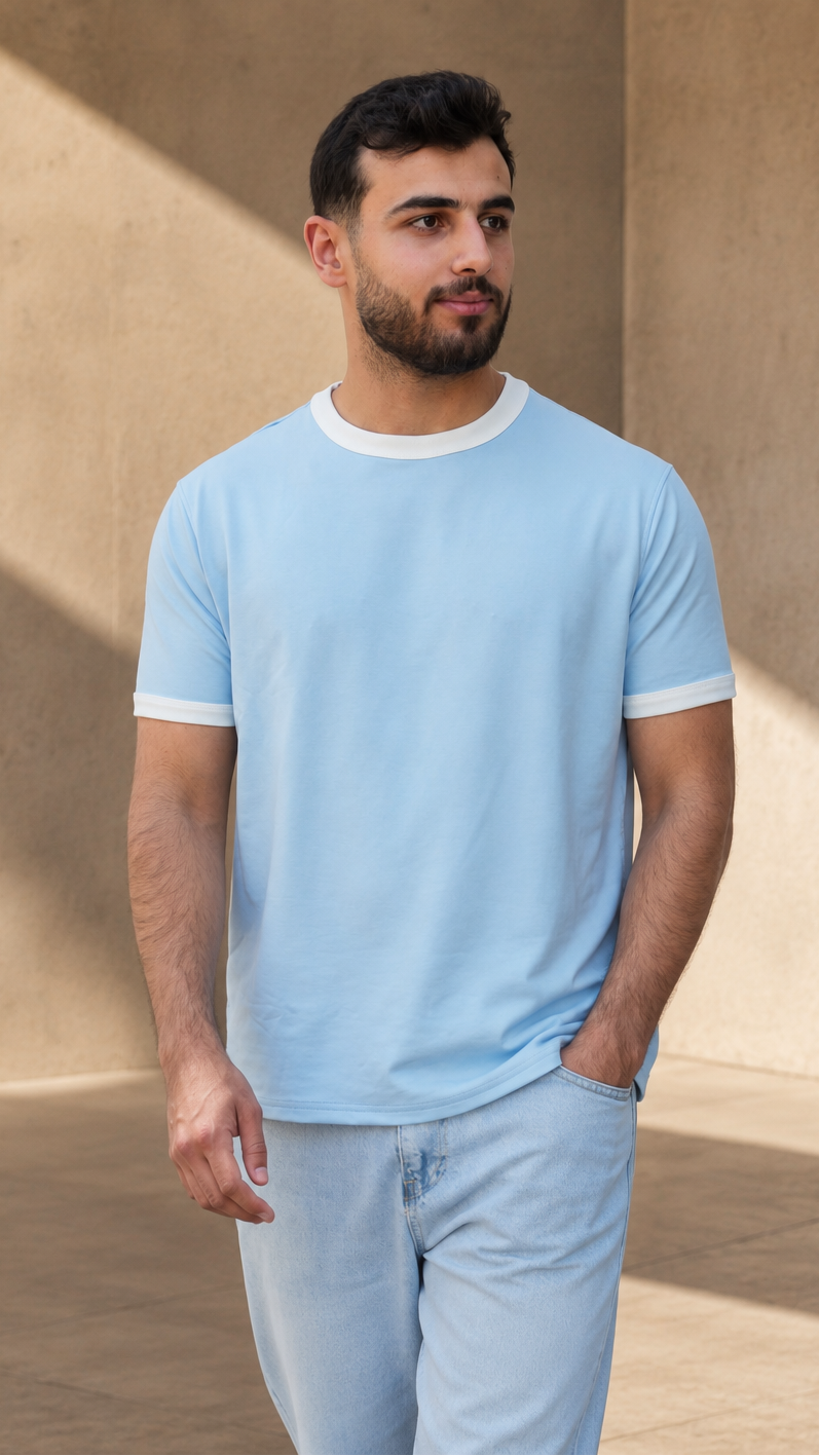 A man wearing a light blue ringer t-shirt with white trim on the collar and cuffs, paired with light blue jeans, standing in a modern architectural setting with warm beige walls and soft shadows.