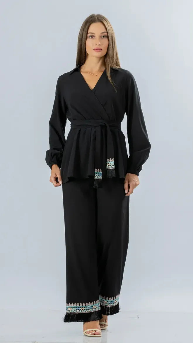 A woman modeling a black long-sleeve wrap-style top and matching wide-leg pants set with small multicolor embroidered details at the hem and waist, photographed against a light gray studio background.