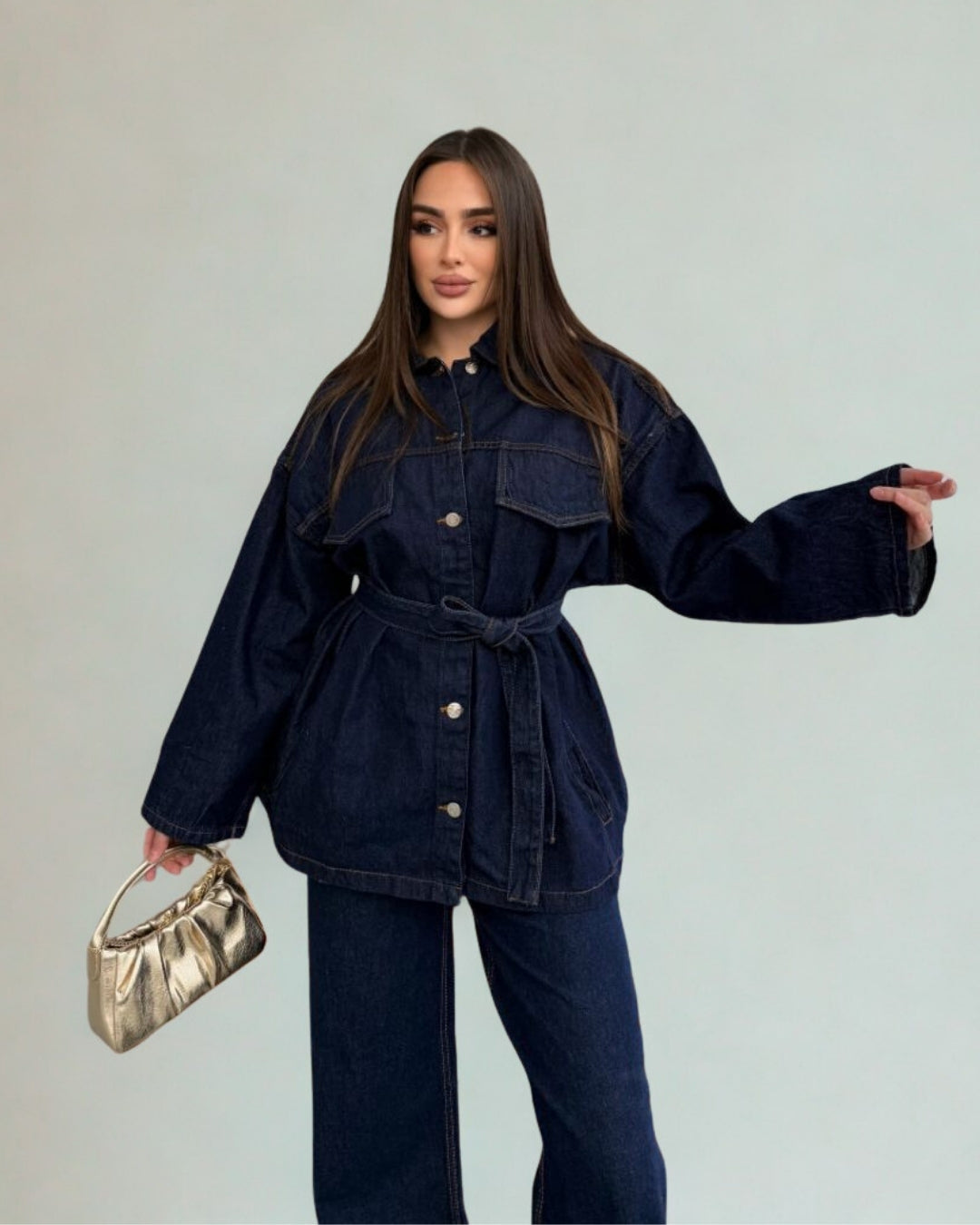 Woman wearing a dark denim jacket and jeans holding a gold handbag against a plain background