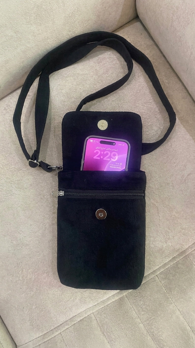 A black velvet phone pouch with a detachable shoulder strap, shown on a light beige fabric surface. A smartphone with a purple screen is tucked inside the pouch.
