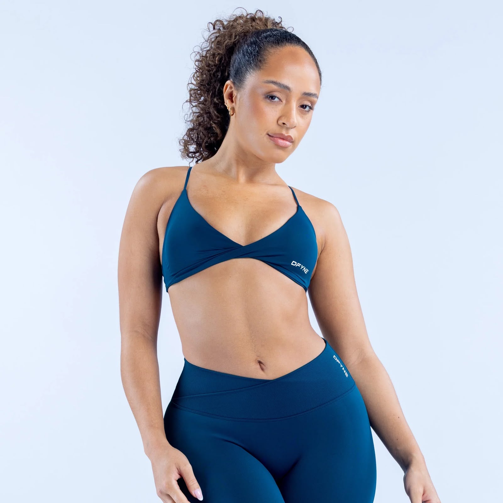 A woman modeling a dark teal blue activewear set consisting of a sports bra and high-waisted leggings, set against a light blue studio background.