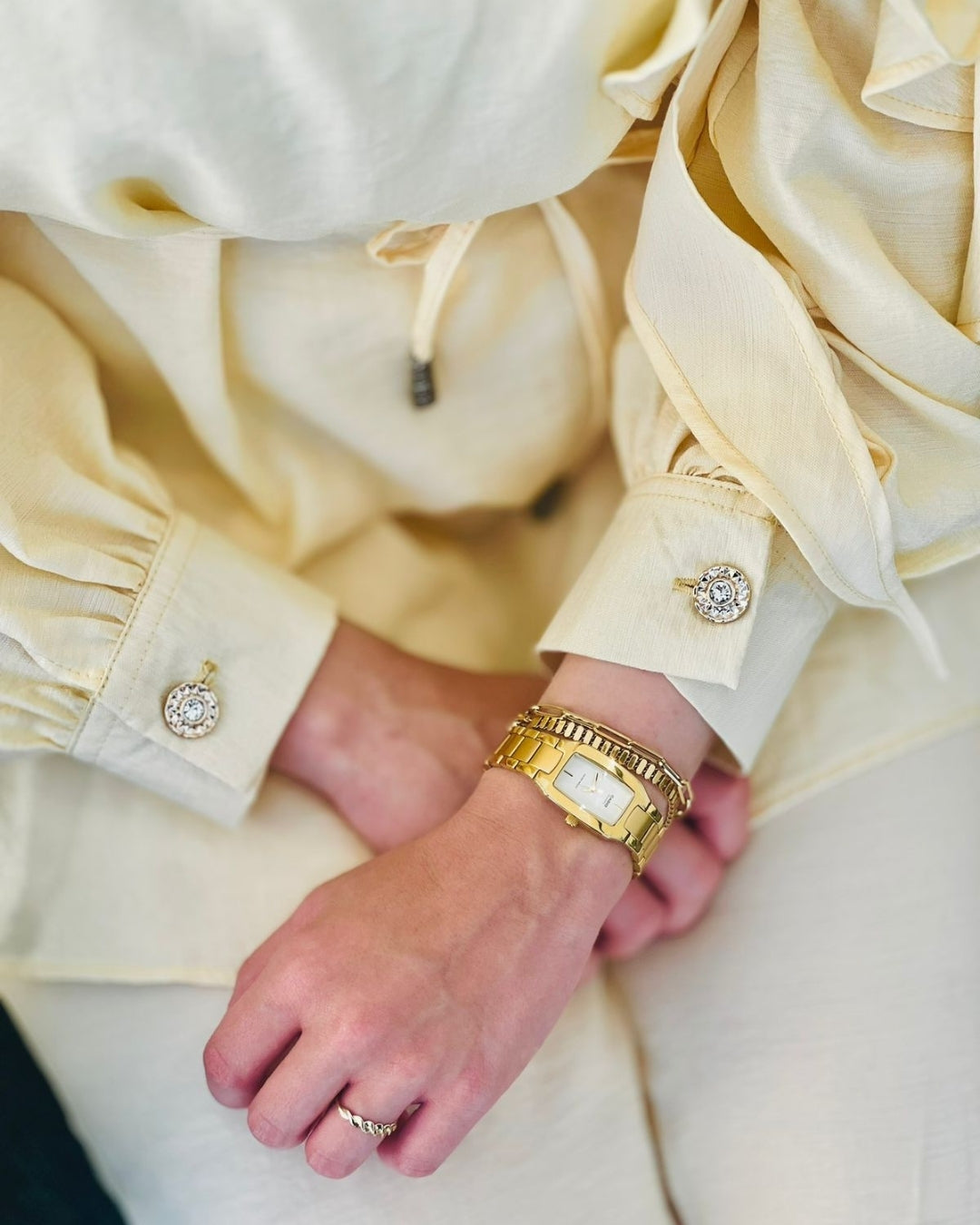 Close-up of a hand wearing a gold watch and rings with a light-colored shirt in the background.