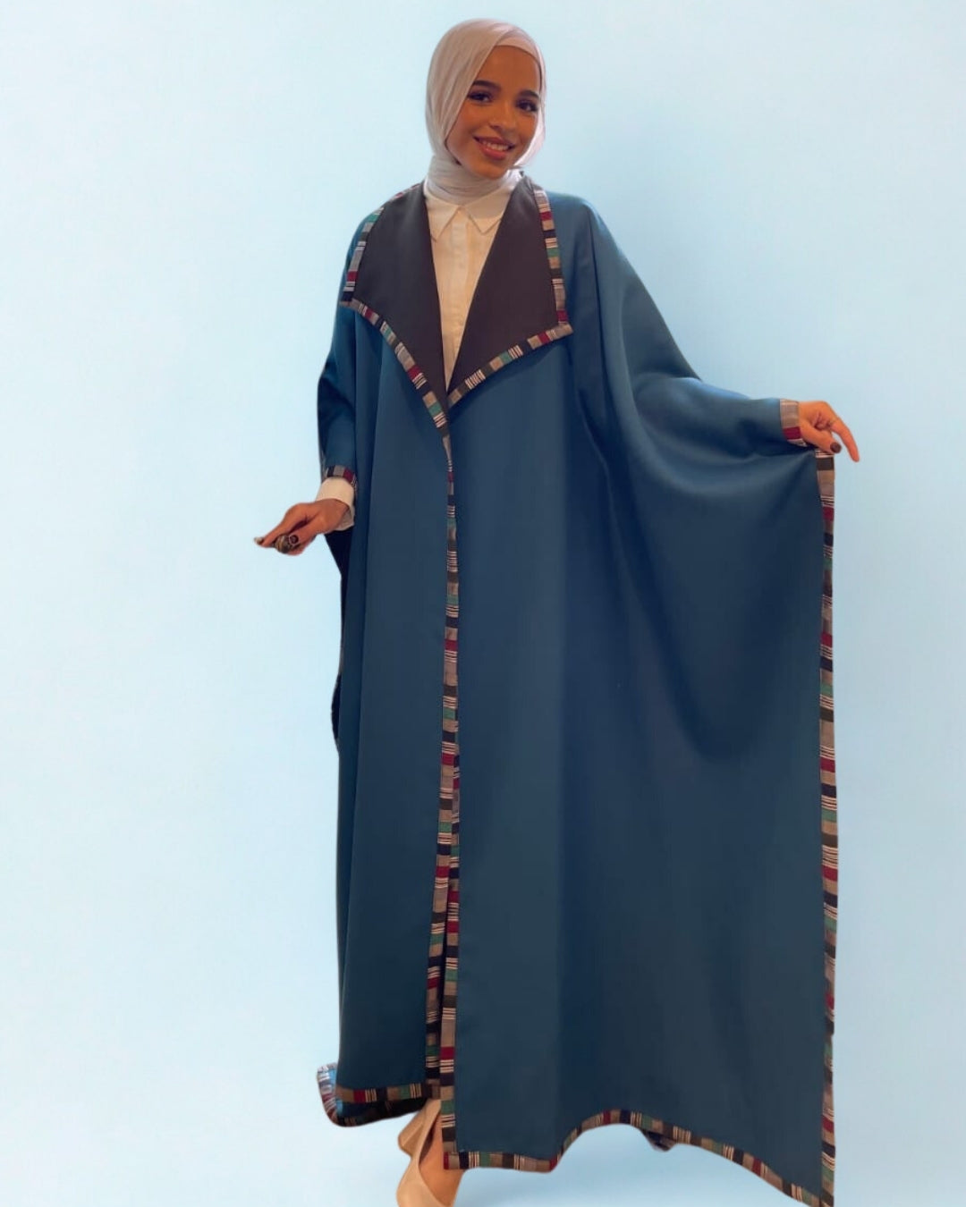 Woman wearing a long, dark blue abaya with patterned trim on a light blue background