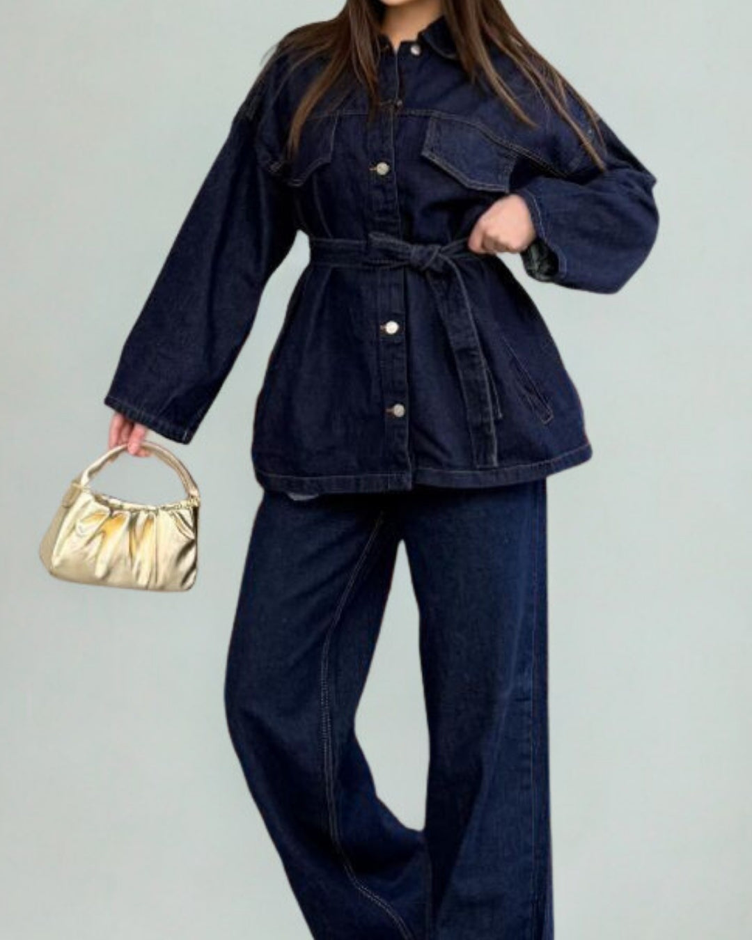 Person wearing a dark denim outfit with a gold handbag on a light gray background