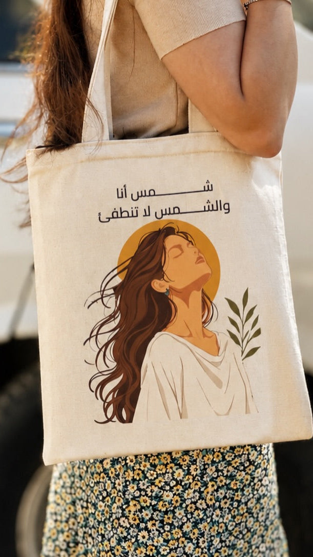 A woman carrying a cream-colored canvas tote bag with Arabic text and an illustration of a woman in a white dress against a yellow halo. The background shows a white vehicle.