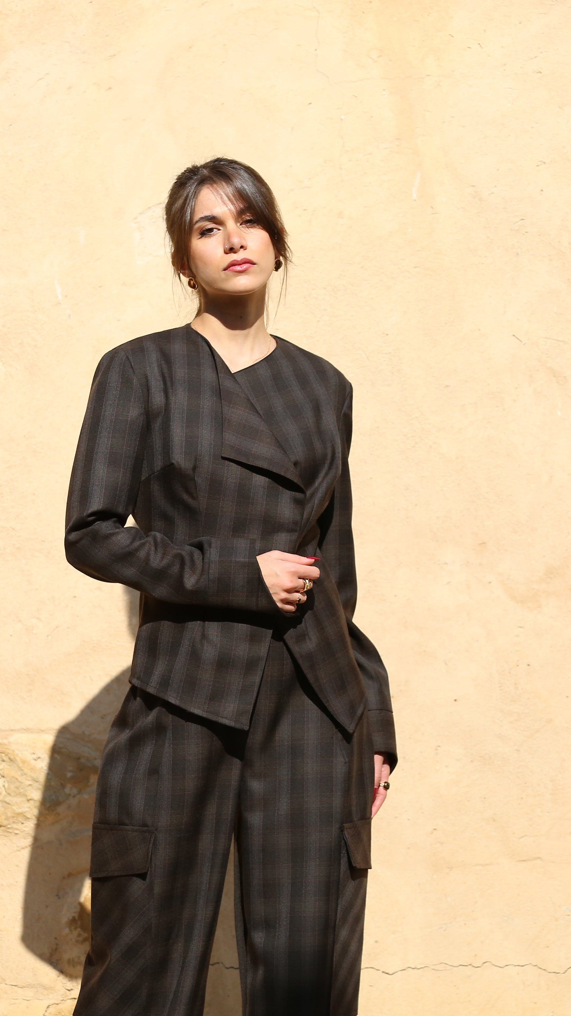 A model wearing a dark gray and black plaid blazer and matching trousers stands against a textured beige stucco wall with a leaf shadow cast on the right.