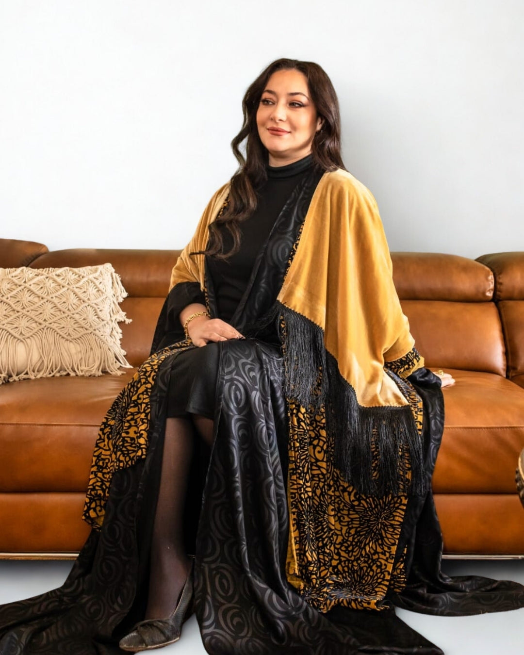 Woman sitting on a brown couch wearing a black and gold patterned shawl.
