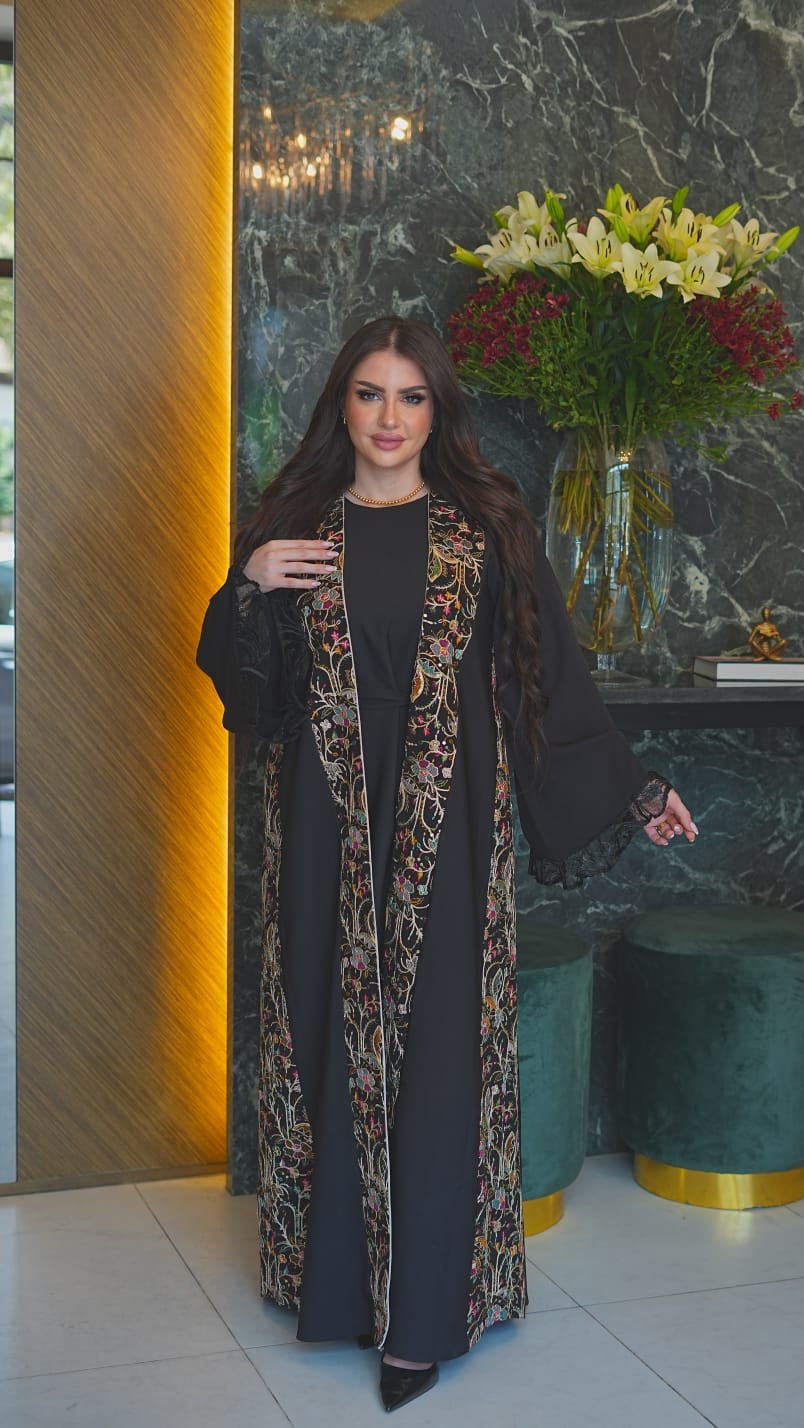 A woman models a long black abaya with a beige and brown patterned outer layer draped over her shoulders, standing in an elegant lobby with a marble wall, floral arrangement, and green velvet stools.