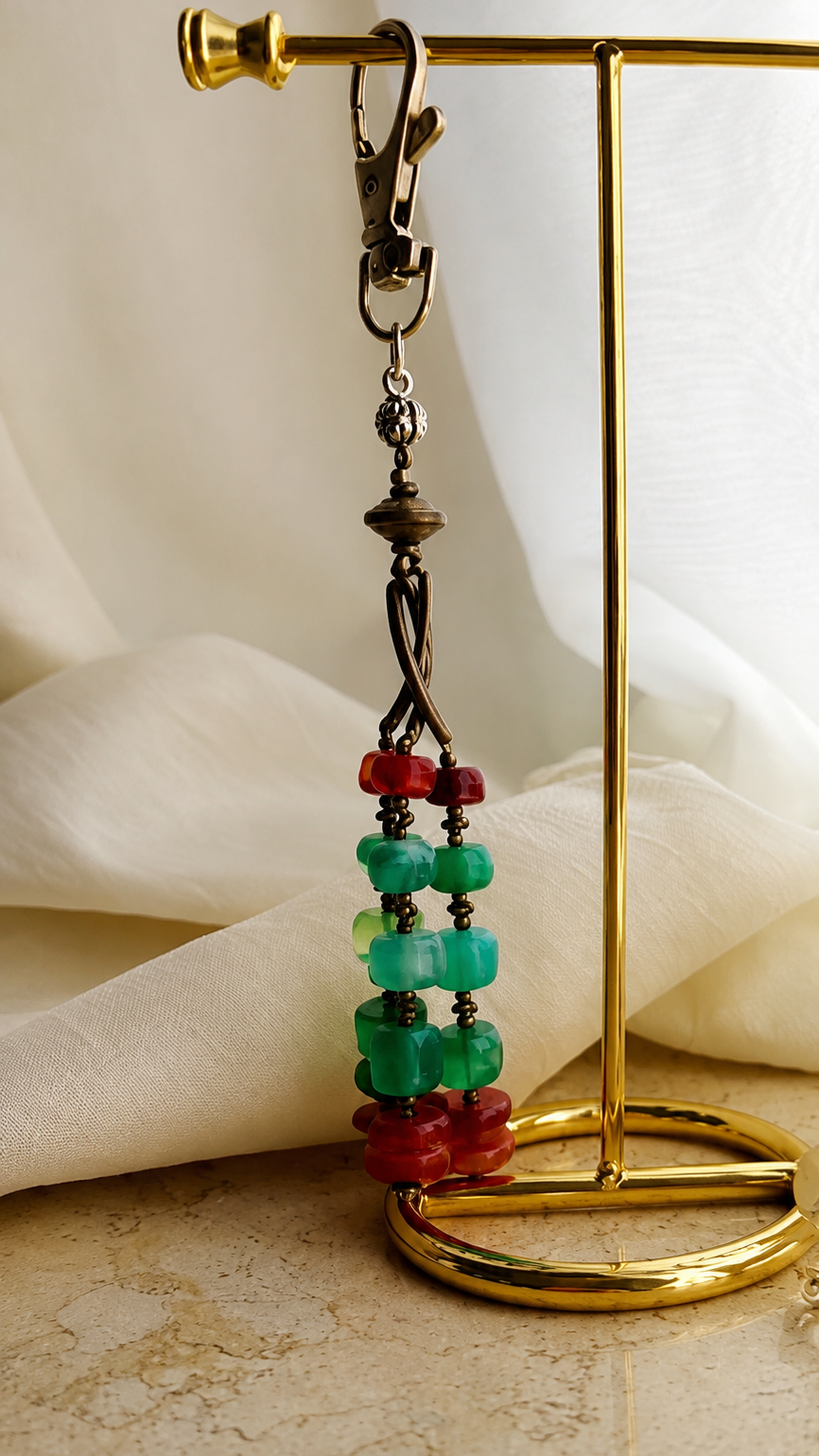 A close-up of a beaded dangle earring with green and red translucent beads, hanging from a gold-tone display stand against a soft cream fabric backdrop.