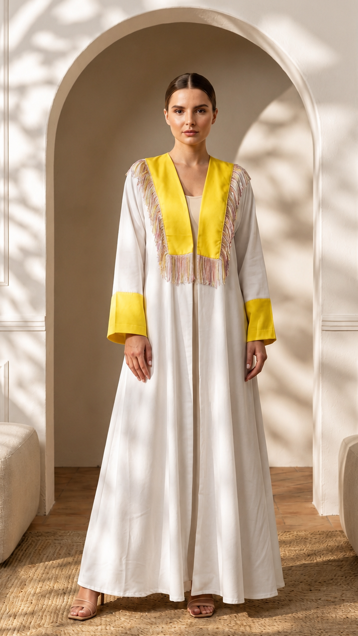 A woman stands in a real interior setting with an arched doorway, wall paneling, floor rug, and upholstered seating, wearing a long white caftan-style dress with bright yellow trim and embroidered details at the neckline and cuffs.