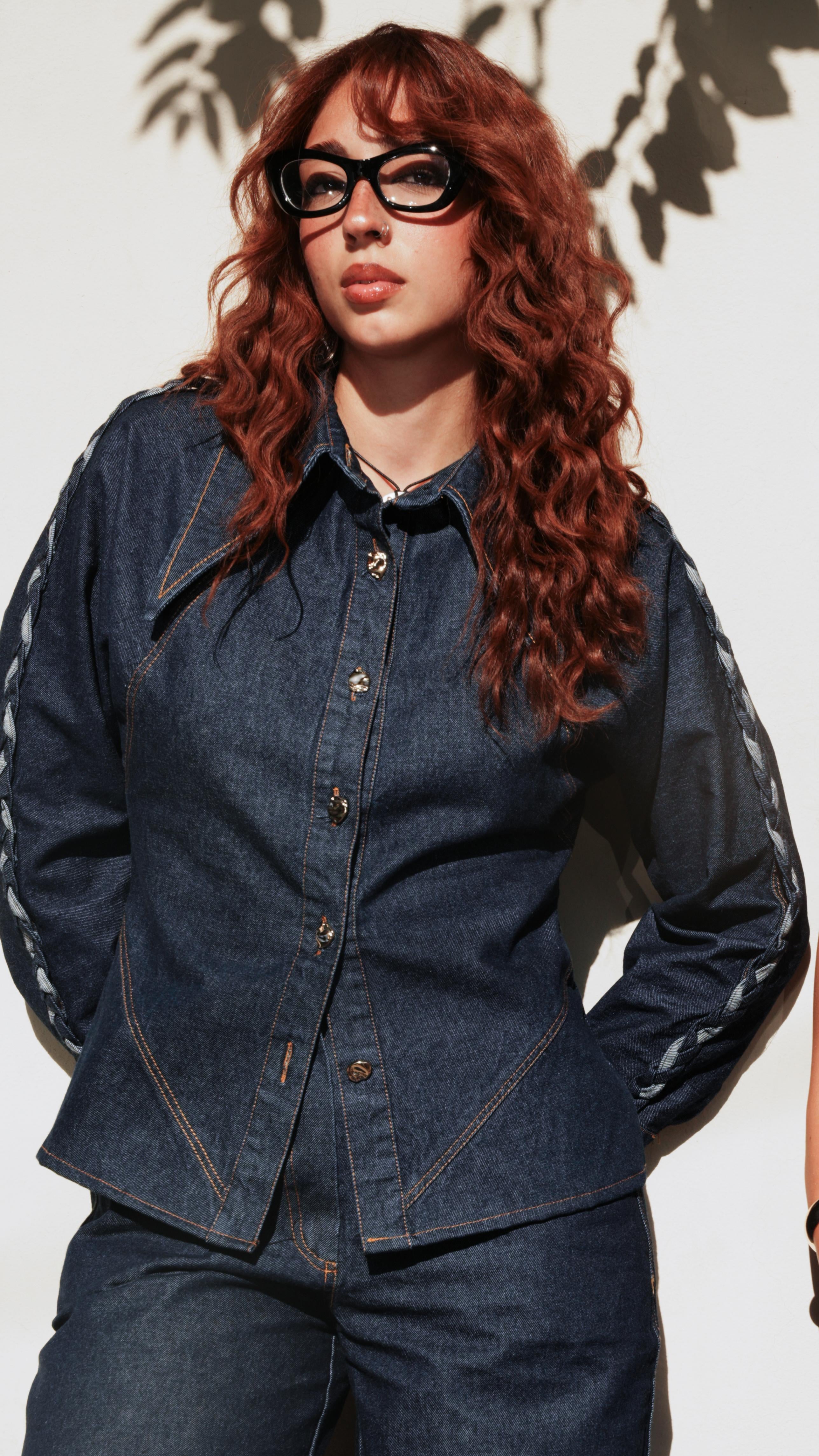 A woman with long wavy red hair and black glasses wearing a dark blue denim jacket with sheer black sleeves and matching dark blue jeans, standing against a white wall with leaf shadows.