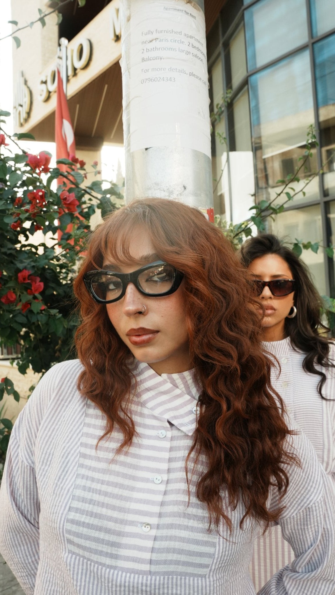 A woman with long, wavy auburn hair and black sunglasses wearing a white and gray striped button-down shirt, standing outdoors in front of a building with a glass facade and red flowers.
