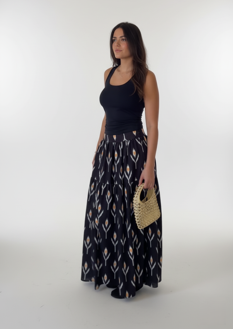 Woman wearing a black sleeveless top and patterned long skirt holding a straw bag on a white background
