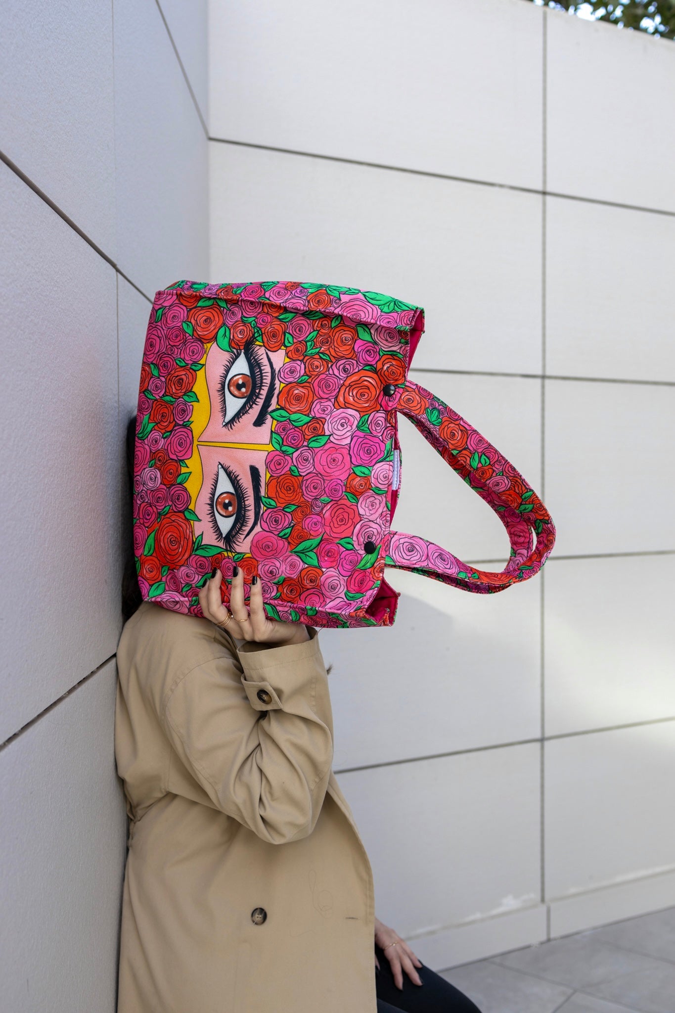 Person holding a colorful bag with floral and face design against a white tiled wall.