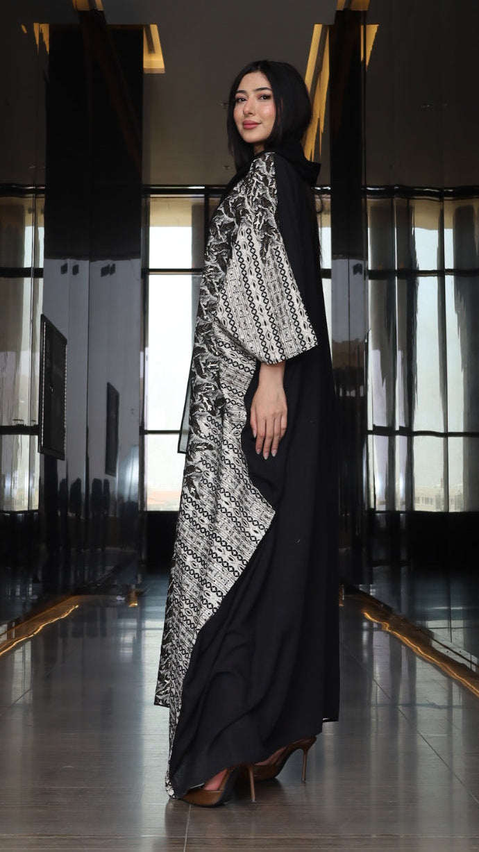 A woman wearing a black abaya with a black and white patterned inner dress and a black hijab, standing in a modern hallway with reflective dark walls.