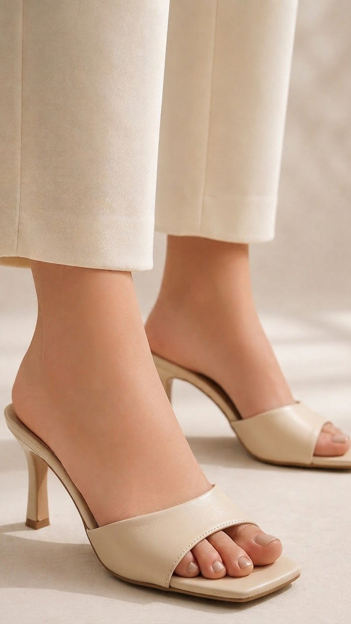 Beige leather mule heels with a square toe and kitten heel, shown on a model wearing cream-colored trousers.
