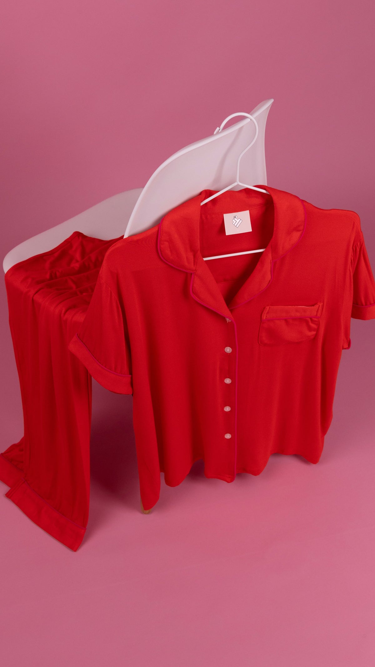 A red short-sleeve button-front shirt with a chest pocket hangs on a white hanger in front of matching red pajama pants draped over a white chair, all set against a solid pink studio background.