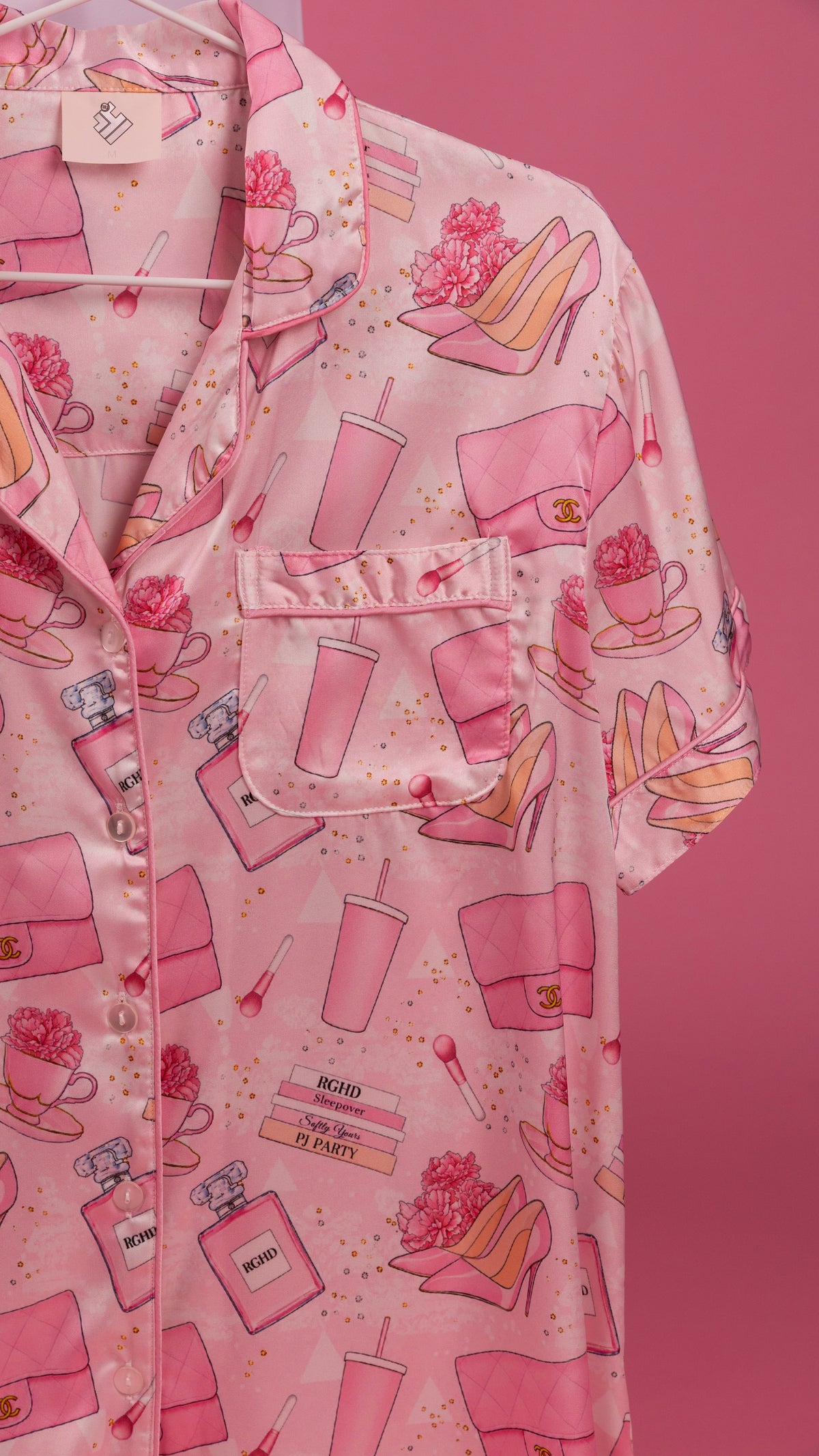 A pink short-sleeve button-up pajama shirt with an all-over print of pink handbags, perfume bottles, high heels, and flowers, shown hanging against a solid pink background.