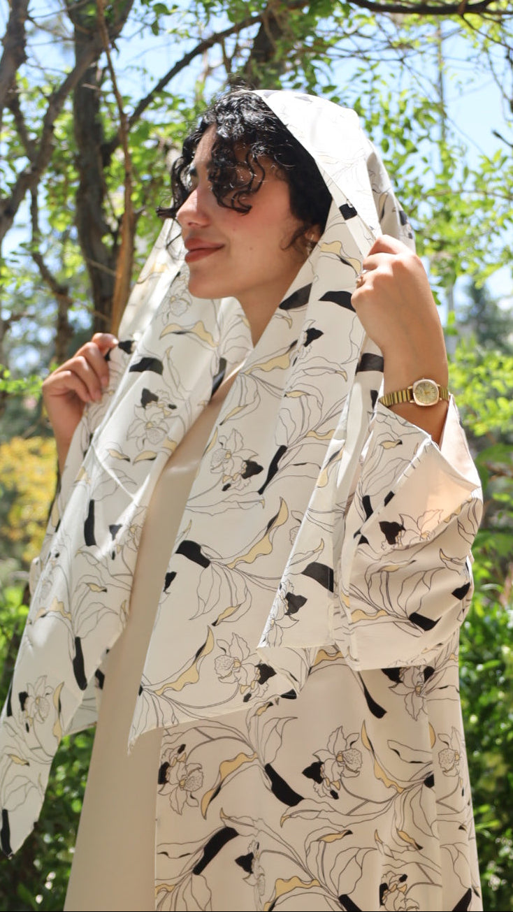 A woman wearing a white hooded robe with a black and gold floral pattern, photographed outdoors under a canopy of green trees and blue sky.