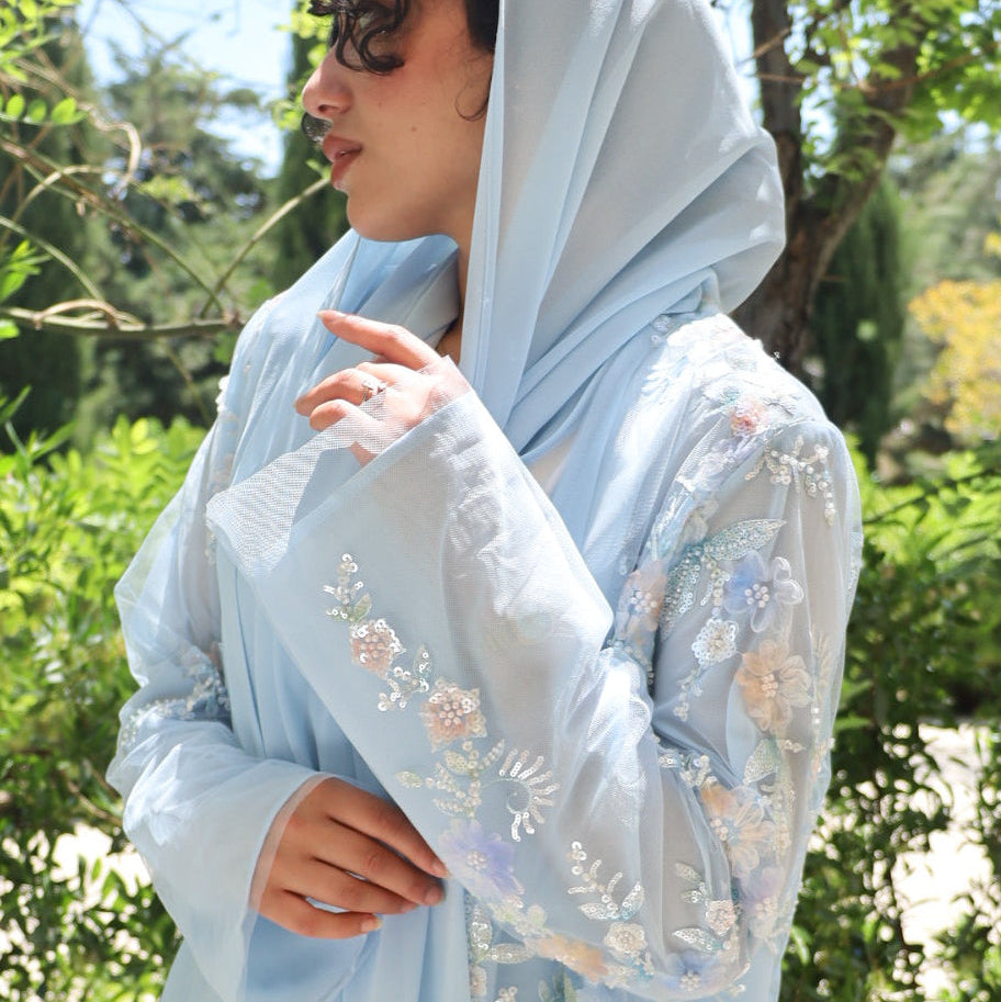A woman wearing a light blue embroidered traditional outfit with a matching headscarf, standing outdoors in front of green trees.