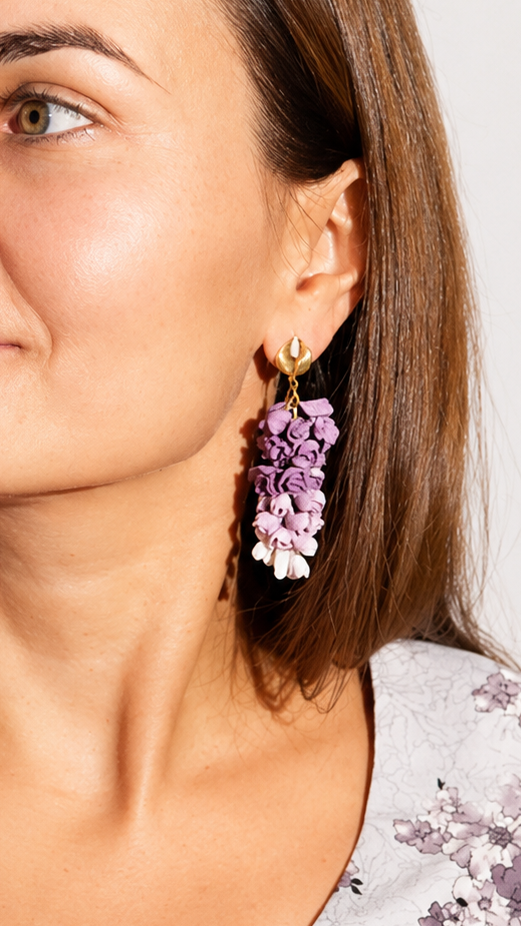 A woman with brown hair is shown in a close-up portrait wearing a light gray floral top and matching purple and white flower-shaped drop earrings.