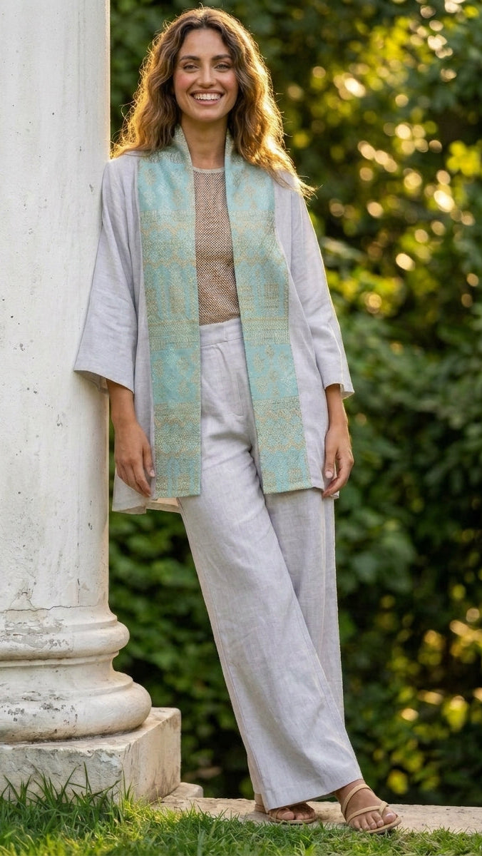 A woman stands outdoors next to a white stone column, wearing a light gray linen outfit with a light blue and gold patterned scarf draped over her shoulders.