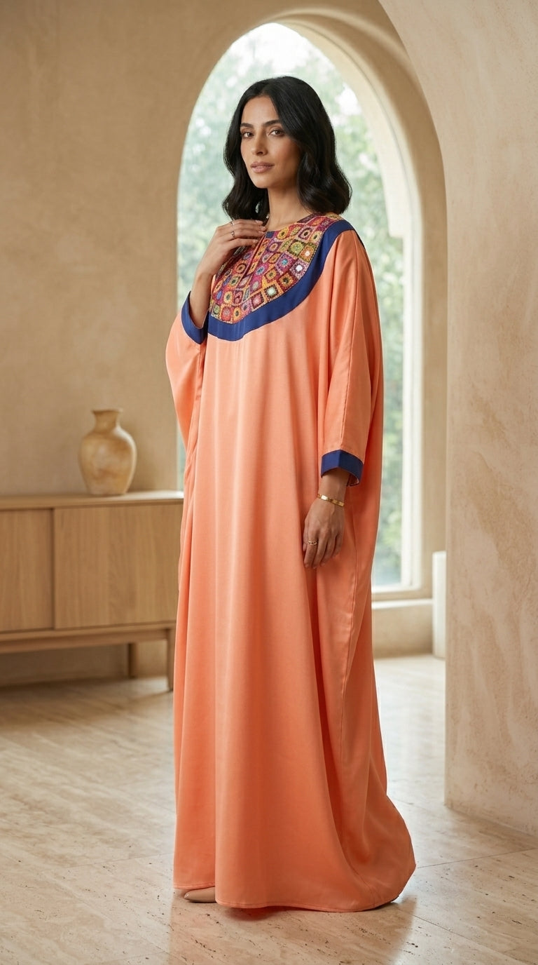 Woman wearing a peach dress with colorful embroidery standing in a room with a wooden cabinet and vase.