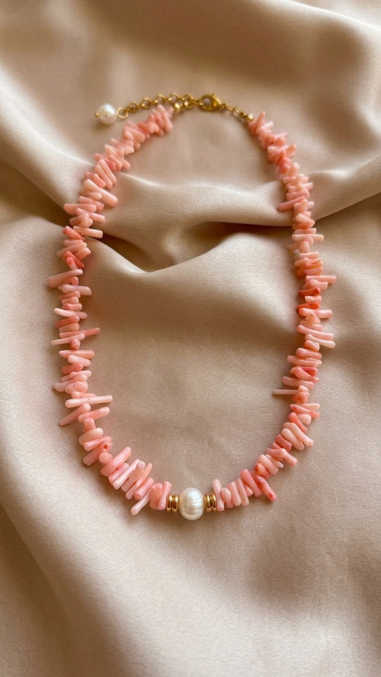 A pink beaded necklace made of irregular shell-like pieces, accented with a central white pearl and gold-tone hardware, displayed on draped beige satin fabric.