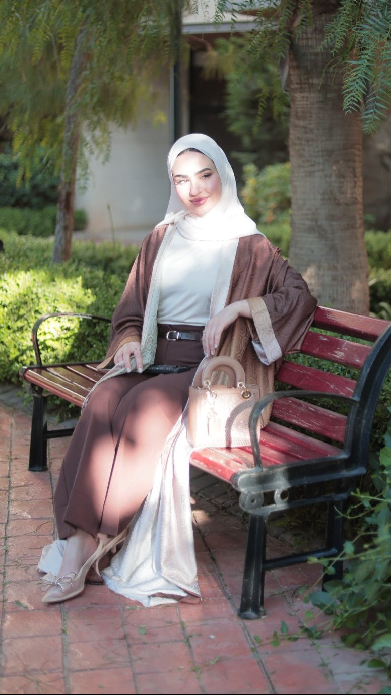 A woman wearing a white hijab, a brown long-sleeved outer garment over a white inner dress, and brown trousers sits on a red wooden park bench. She is holding a beige handbag in a garden setting with trees and greenery.