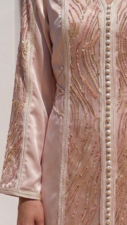 A woman wearing a light pink satin abaya dress with intricate gold embroidery and beadwork, paired with a white hijab, standing in front of a white arched wall.