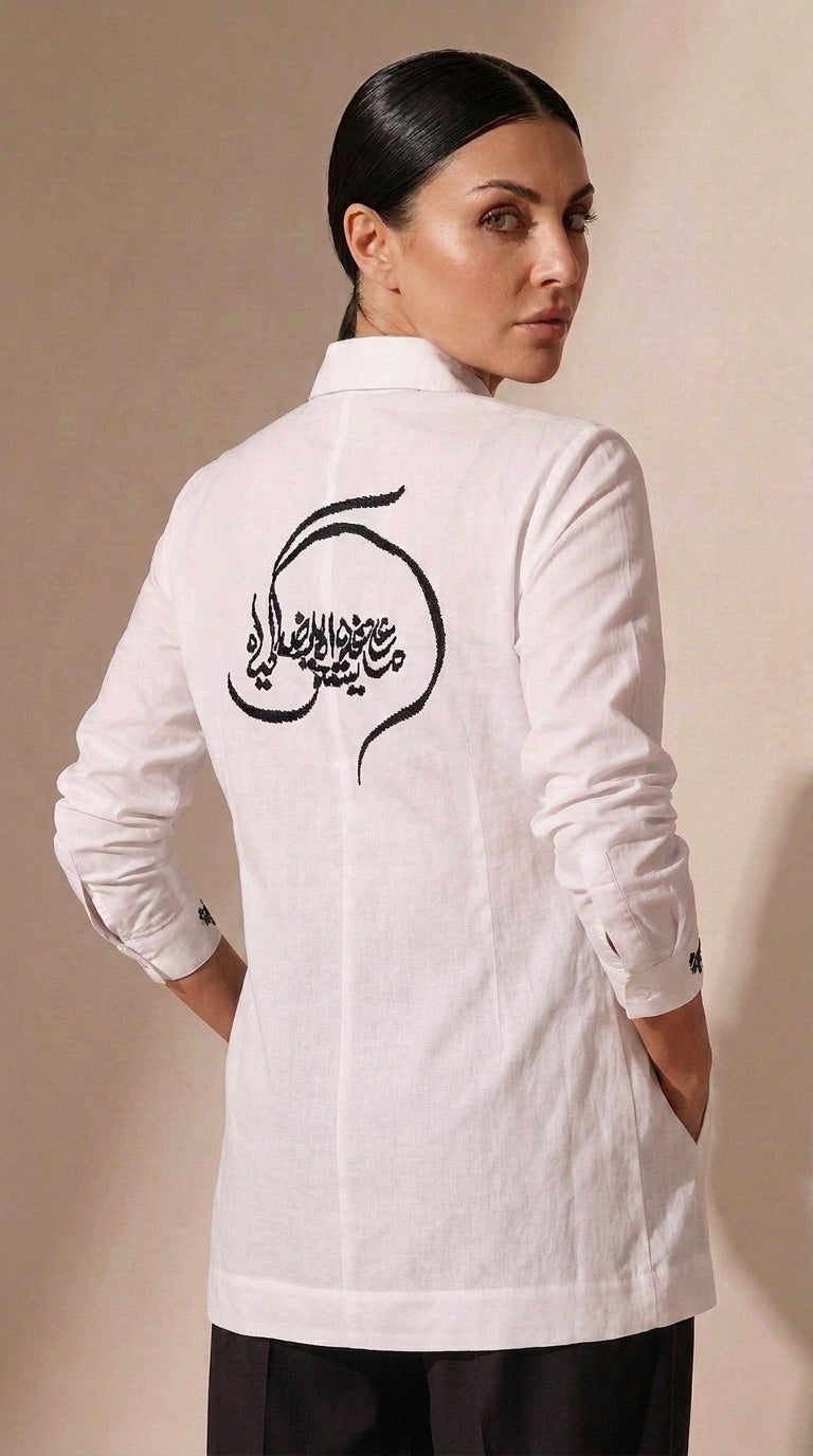 A woman shown from the back wearing a white long-sleeve tunic-style top with black Arabic calligraphy printed across the upper back, paired with dark pants, against a plain beige studio background.