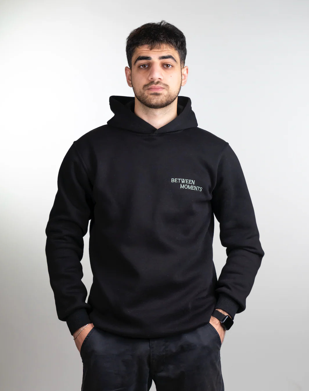 Person wearing a black hoodie with 'Between Moments' logo on a plain background