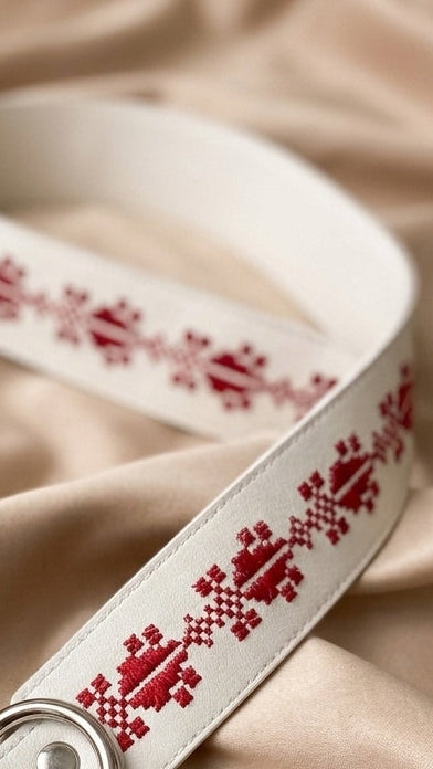A white belt with a red embroidered traditional pattern, draped on a beige satin fabric background.