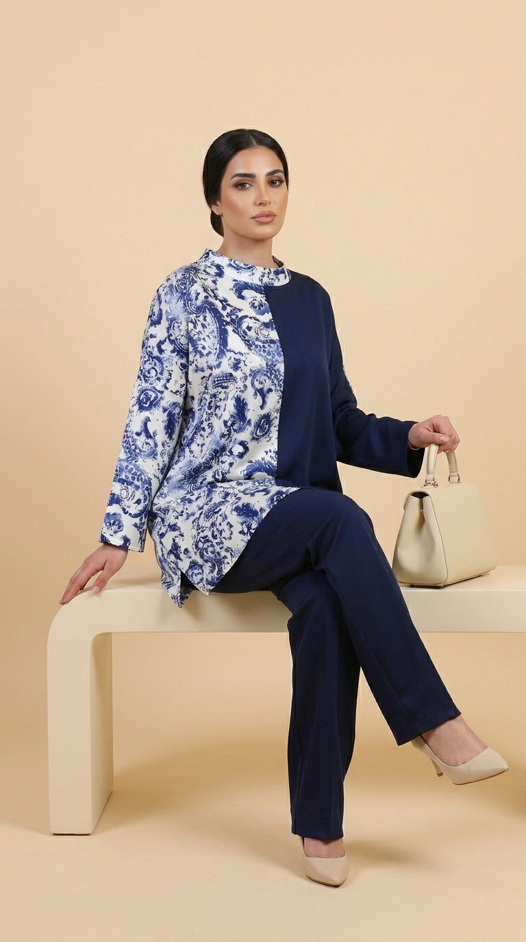 Woman sitting on a bench wearing a blue and white floral top with navy pants against a beige background