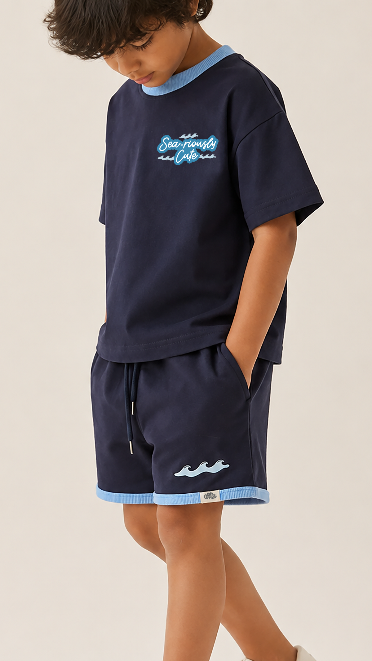 A child wearing a navy blue short-sleeve t-shirt and matching shorts set with light blue trim and graphic details, standing against a plain light beige background.