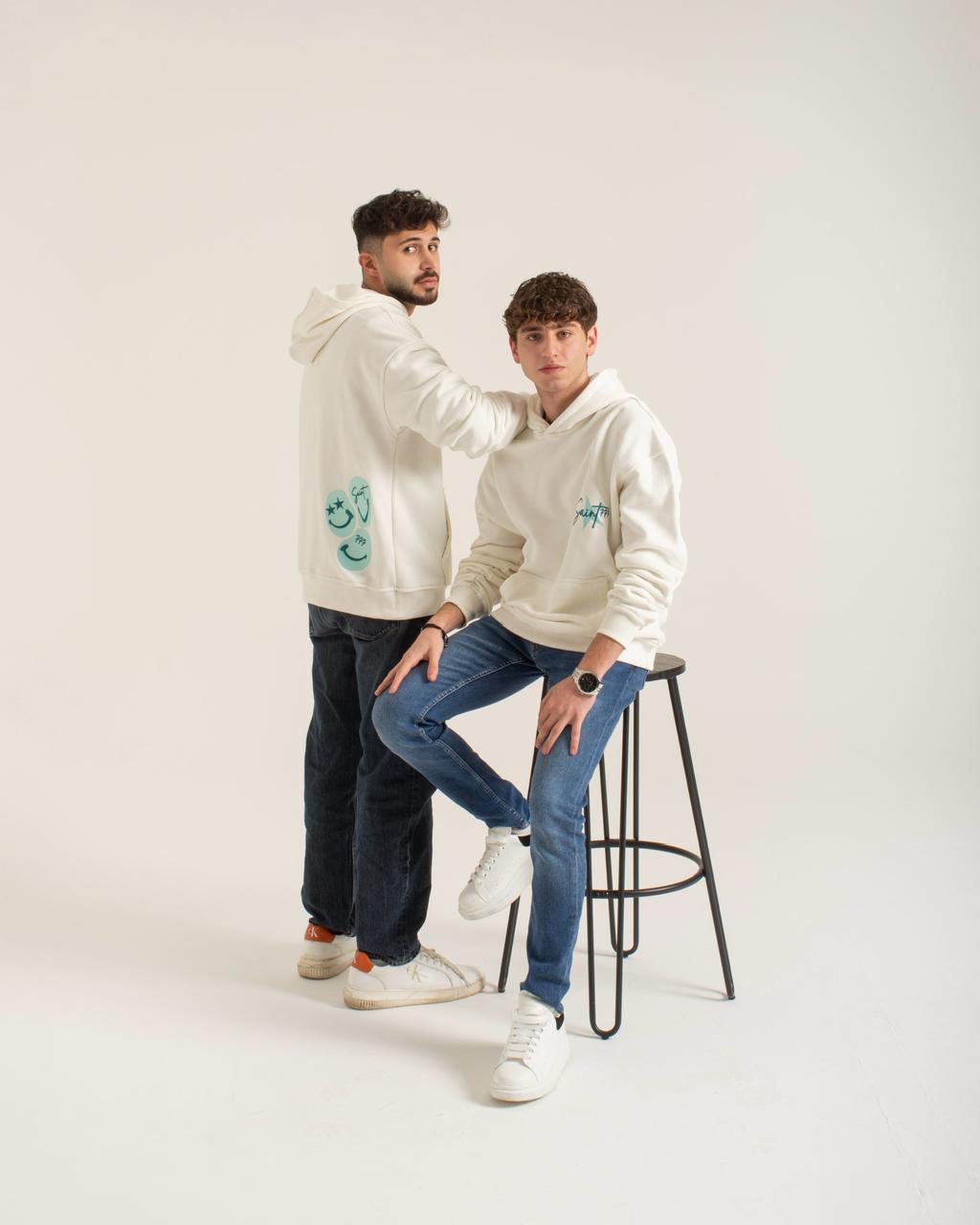 Two people wearing white hoodies with blue designs on a plain background