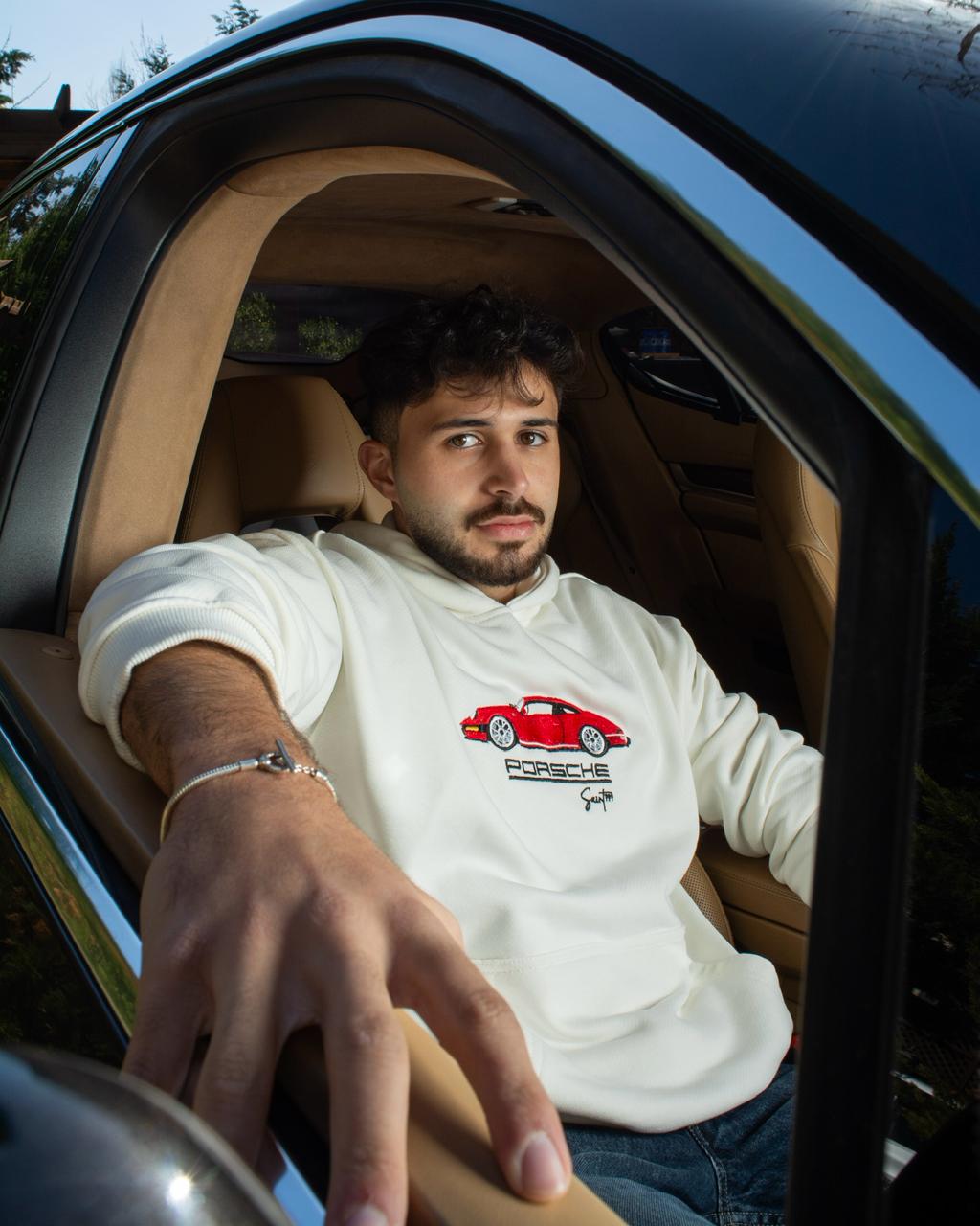 Man sitting inside a car wearing a white Porsche t-shirt with a red car graphic.