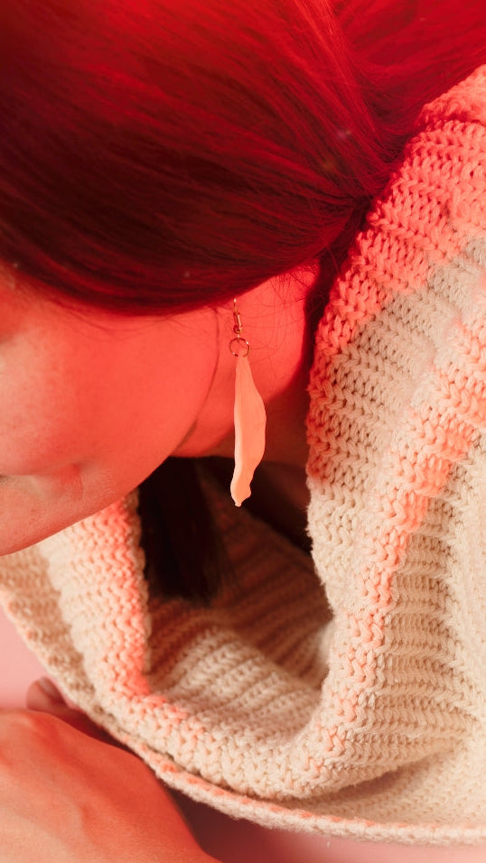 A close-up lifestyle shot of a woman with red hair wearing a chunky knit cowl neck warmer in cream and pink stripes, under warm red ambient lighting.