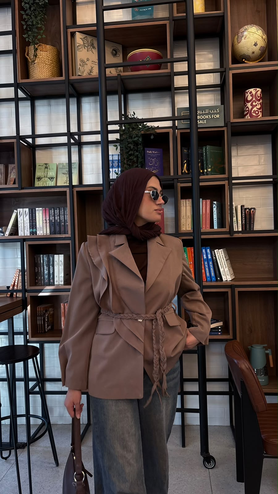 A woman wearing a brown double-breasted belted blazer, dark brown turtleneck, blue jeans, and a dark brown hijab stands in front of a large black metal bookshelf filled with books and decor.