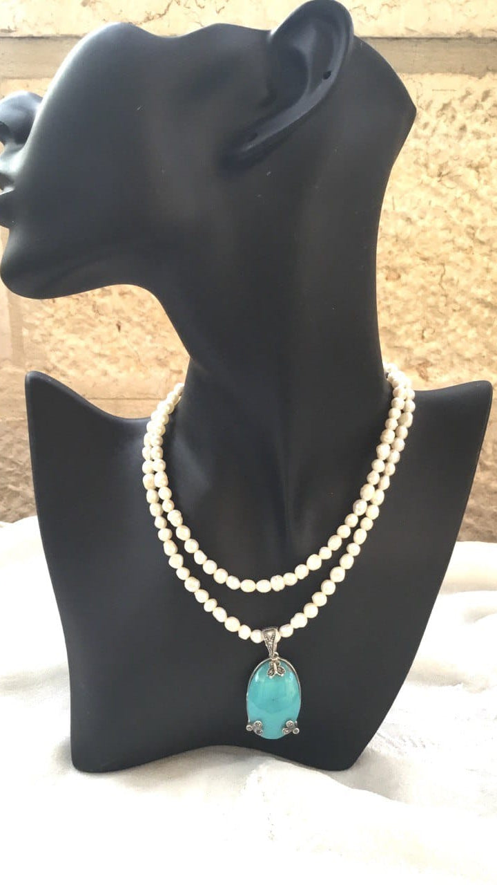 A double-strand pearl necklace with a turquoise pendant, displayed on a black mannequin bust against a textured beige stone wall background.