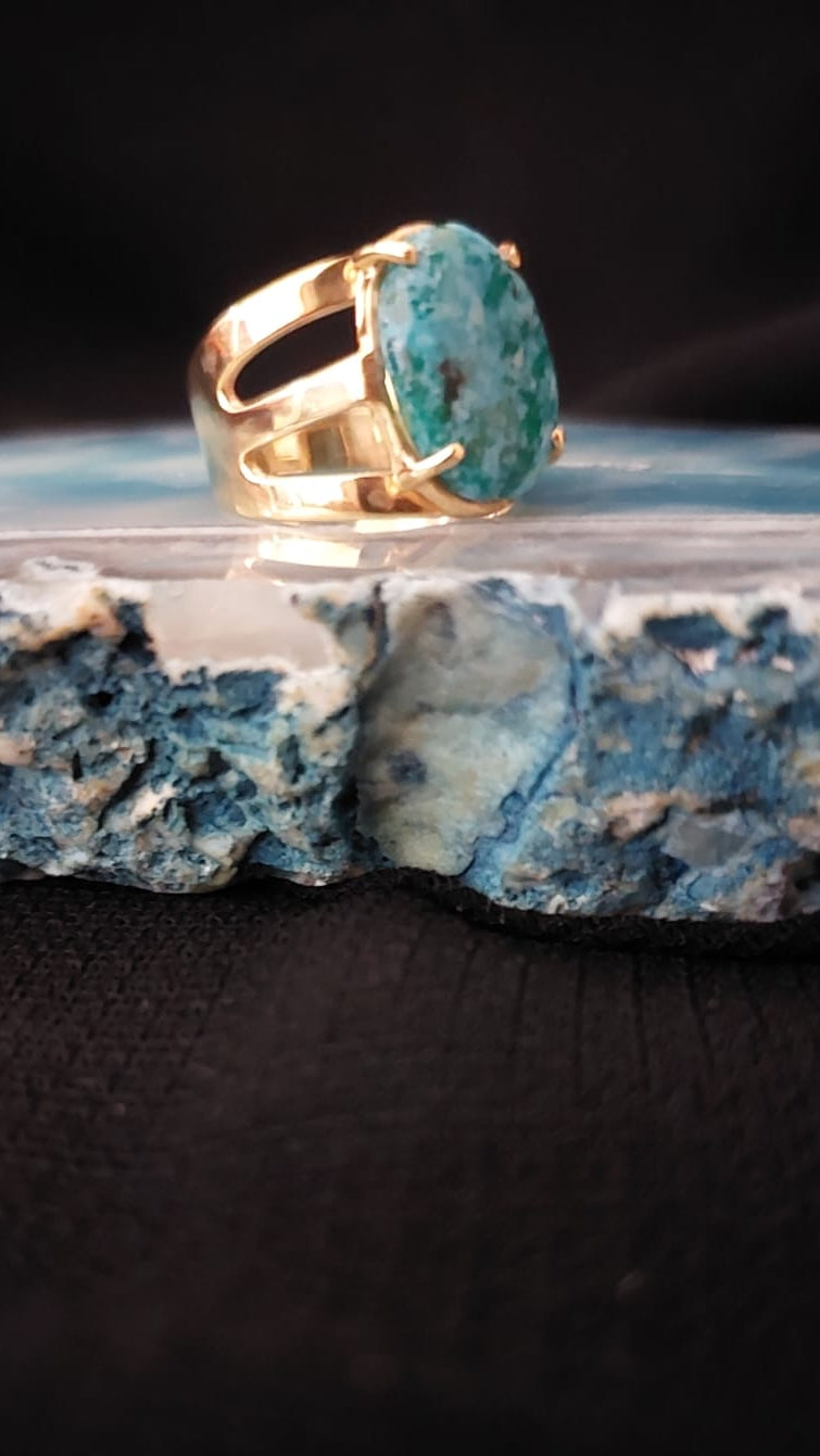 A gold ring with a large oval turquoise-blue stone, displayed on a polished blue and white mineral slab against a black background.