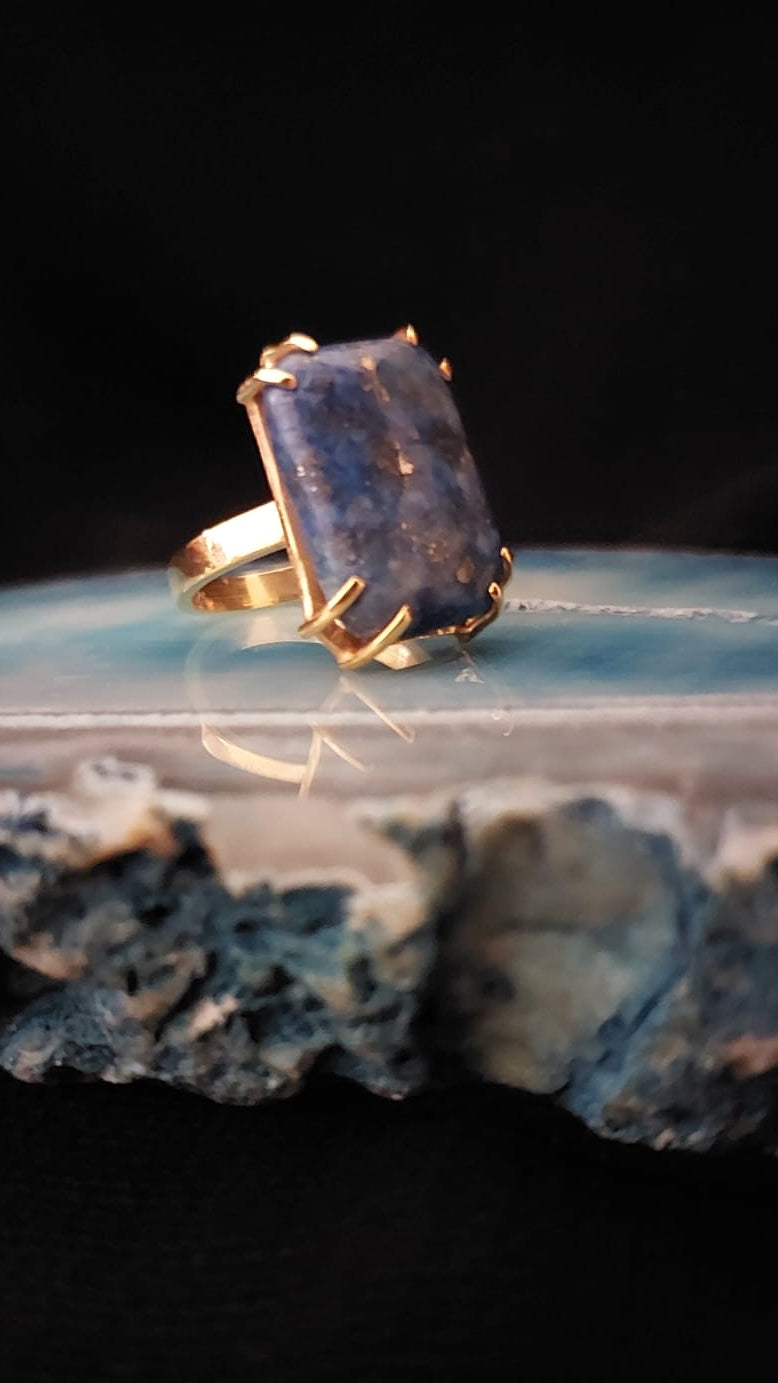 A gold ring with a rectangular dark blue stone, resting on a polished blue agate slice against a black background.
