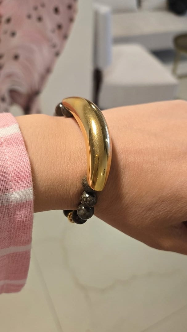 A person's wrist wearing a chunky, polished gold cuff bracelet with a black beaded accent strand underneath, set against a blurred indoor background.