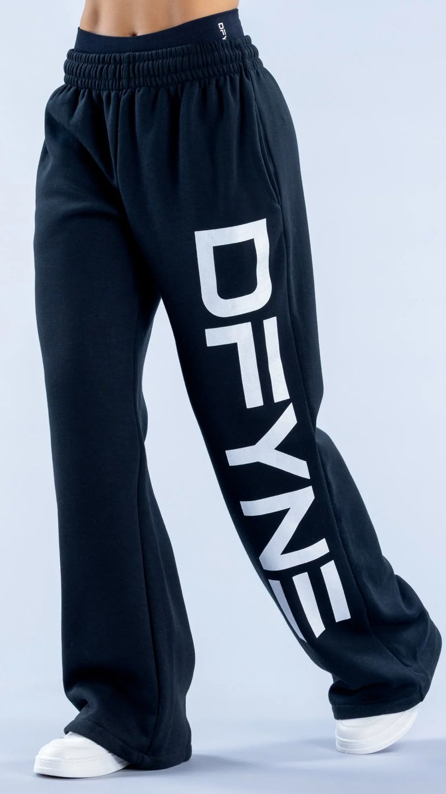 Black wide-leg sweatpants with a large white vertical 'DFYNE' logo printed on the left leg, shown on a model from the waist down against a light gray studio background.