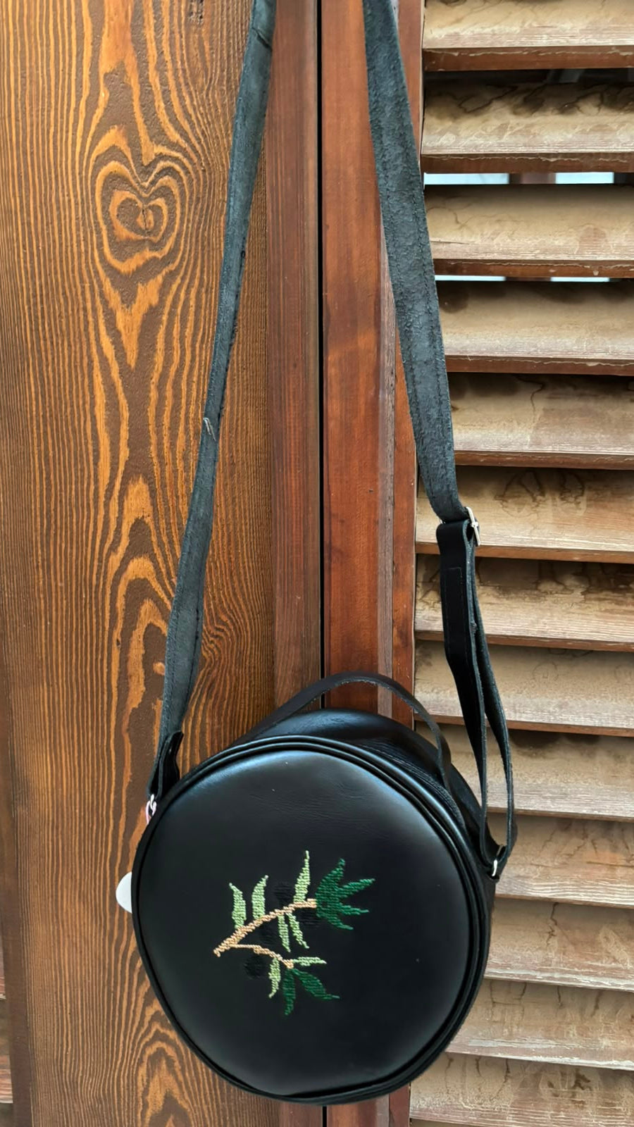 A black round shoulder bag with a green embroidered leaf branch design, hanging from a dark strap against a wooden slatted background.