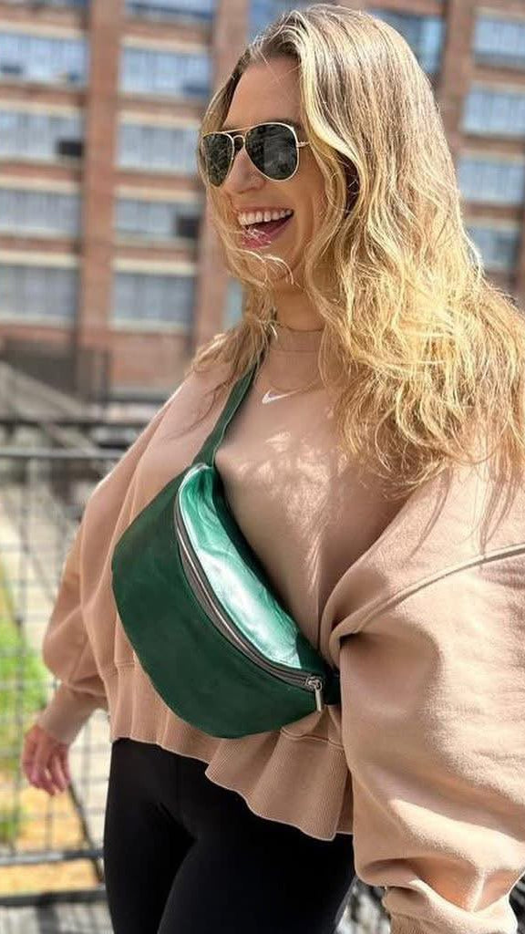 A woman wearing aviator sunglasses, a beige sweatshirt, and black leggings is pictured outdoors on a balcony, wearing a dark green crescent-shaped belt bag with a shiny mint green interior. A brick building is visible in the background.