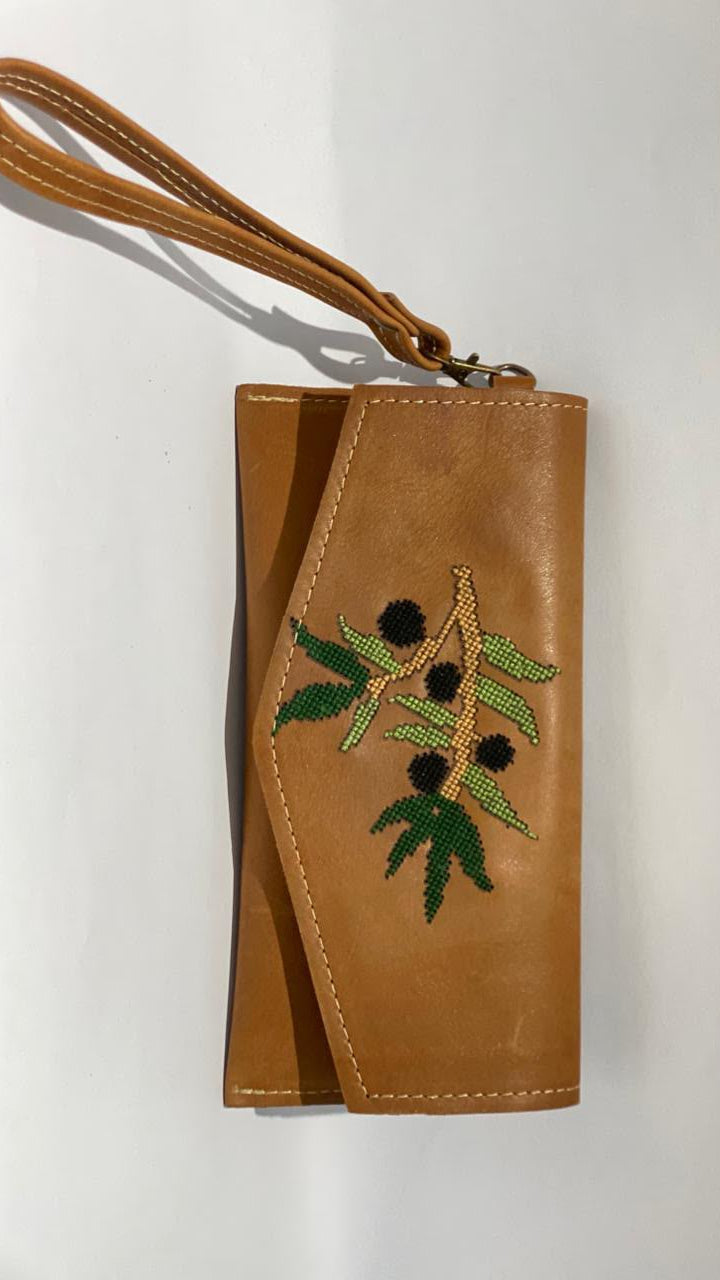 A tan leather wristlet clutch with a matching wrist strap, featuring an embroidered olive branch design with green leaves and black olives on the front flap, set against a plain white background.