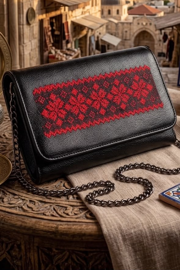 A black leather shoulder bag with a red floral embroidered pattern across the front flap, resting on a carved wooden table with a beige cloth and a metal chain strap. The background shows a scenic view of Jerusalem's old city with domes and arches.