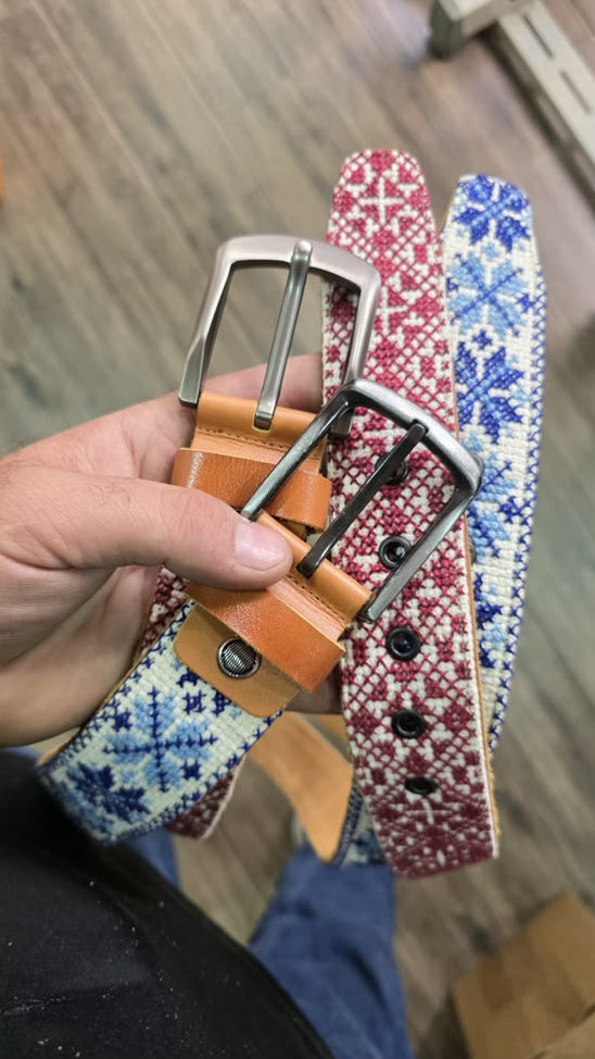 A hand holding three woven belts with silver buckles. The belts feature a tan leather base with blue and white geometric patterns on one, a red and white pattern on another, and a tan leather belt with a silver buckle on the third.