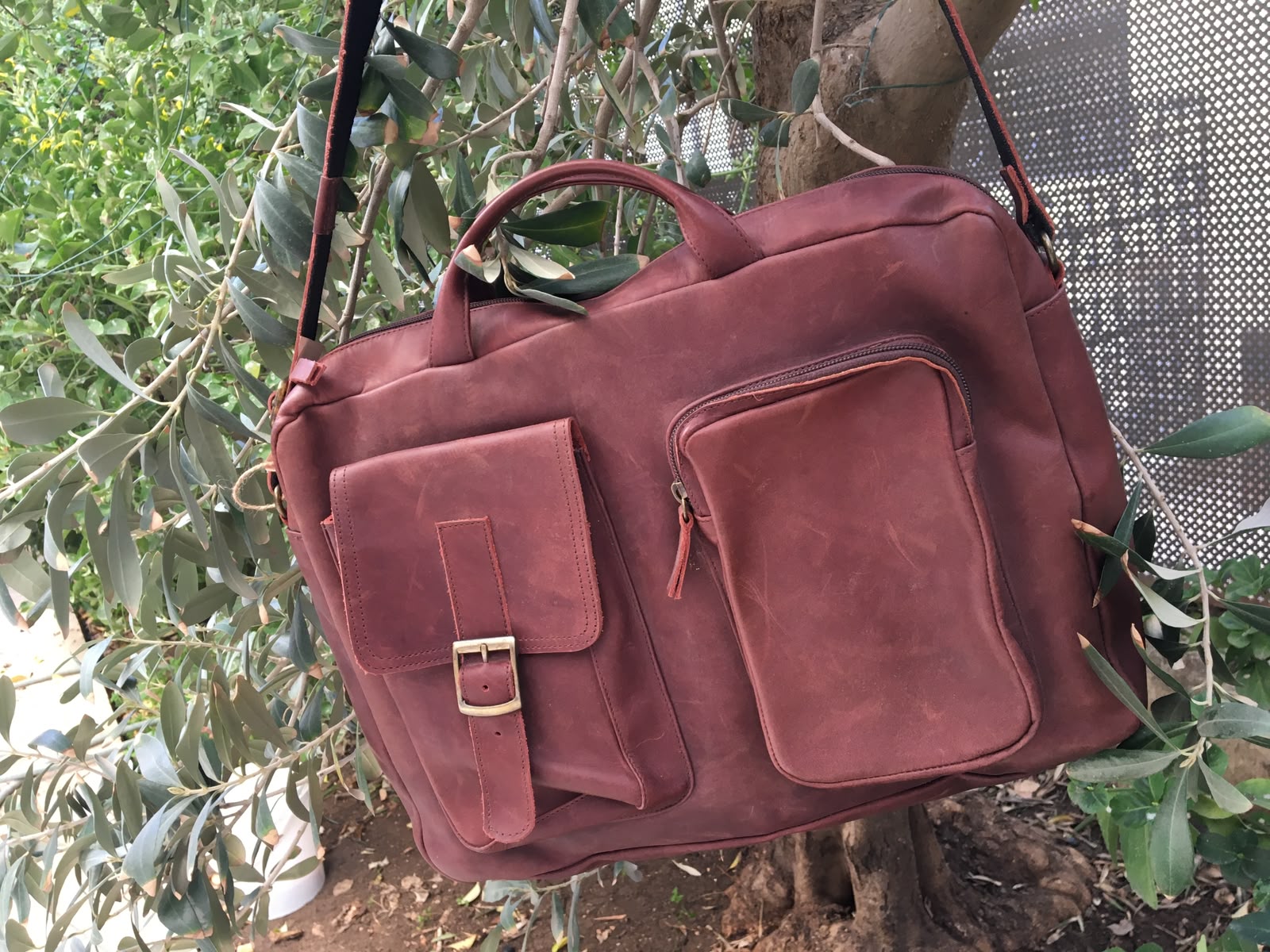 A reddish-brown leather briefcase or laptop bag with dual top handles, a front flap pocket with a buckle, and a zippered side pocket, hanging outdoors against a backdrop of green leaves and tree branches.